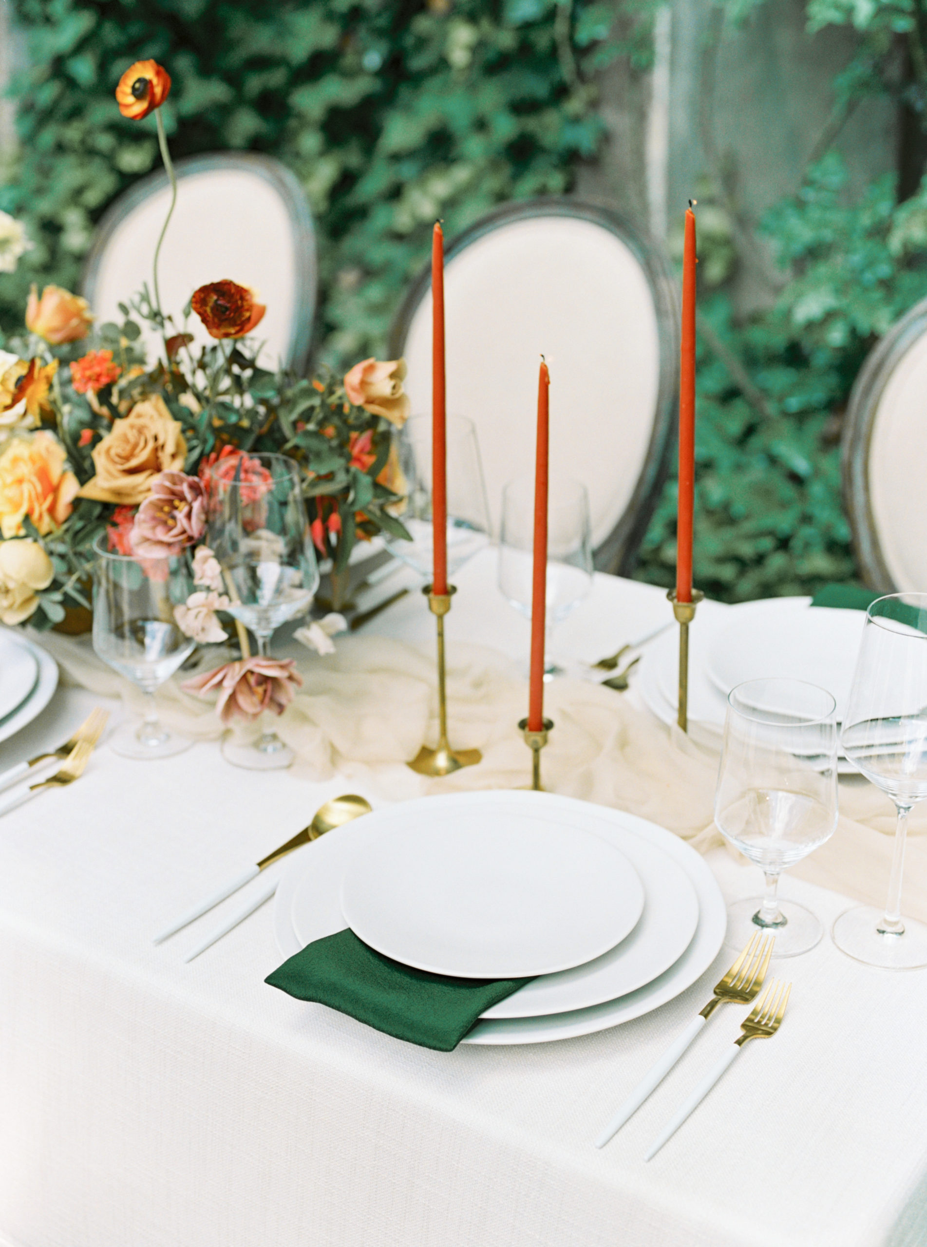 Thin, orange candle sticks on a white table scape with emerald napkins and gold and white flatware