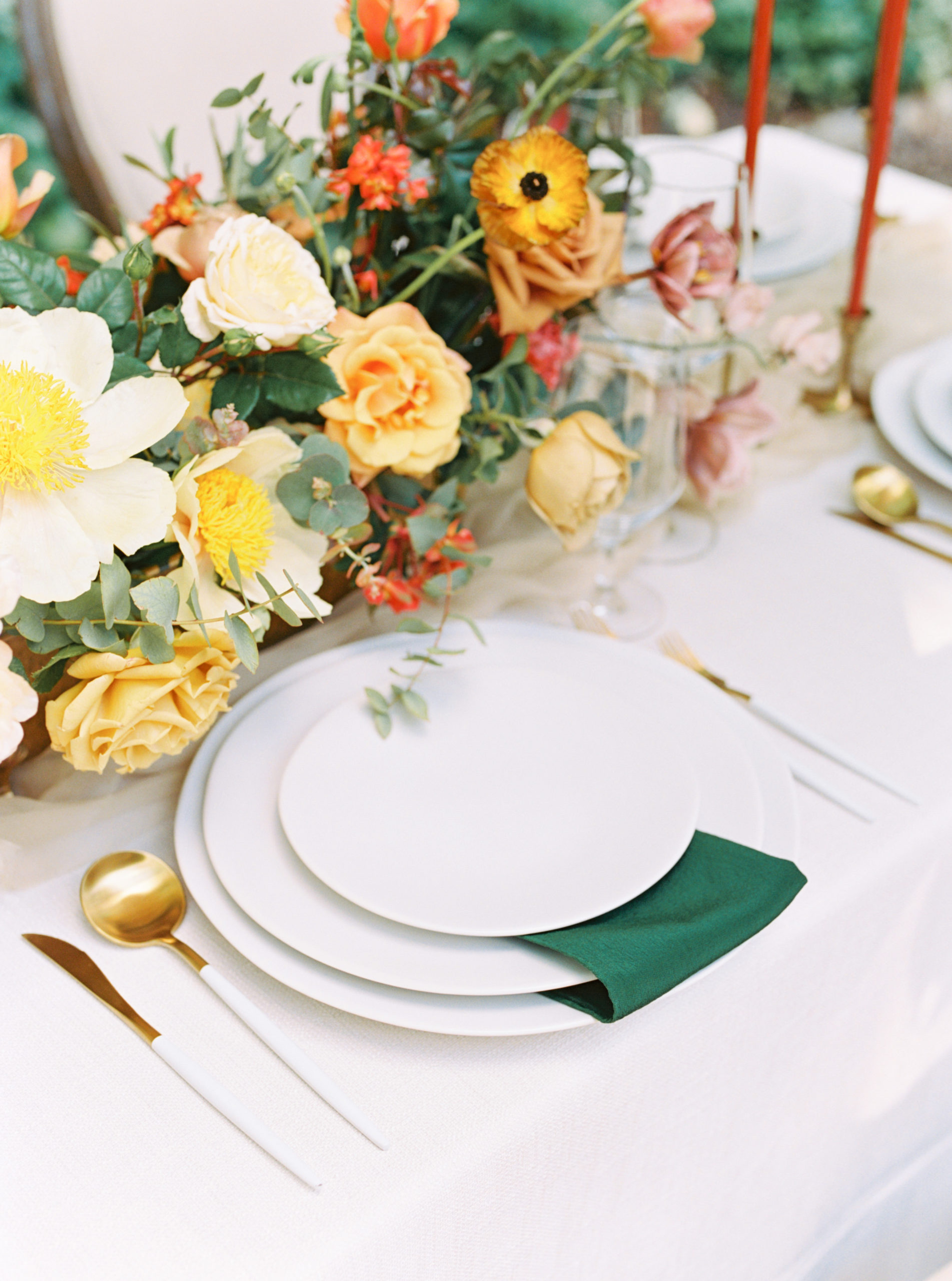 White layered place setting with emerald napkin and orange and yellow florals and gold flatware
