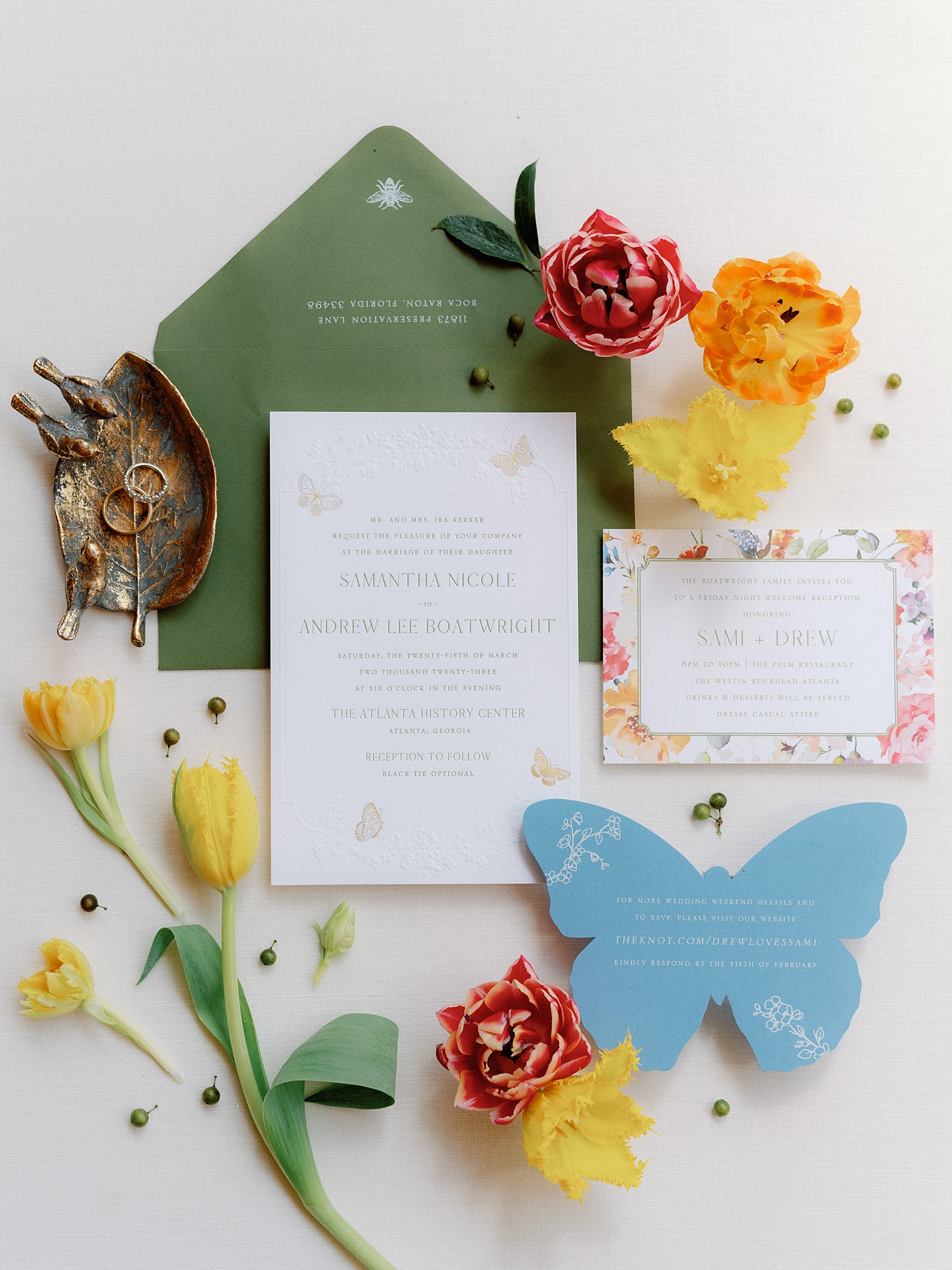 A flat lay of stationery that includes a white invitation, green envelope and blue butterfly info card. All are adorned with flowers.