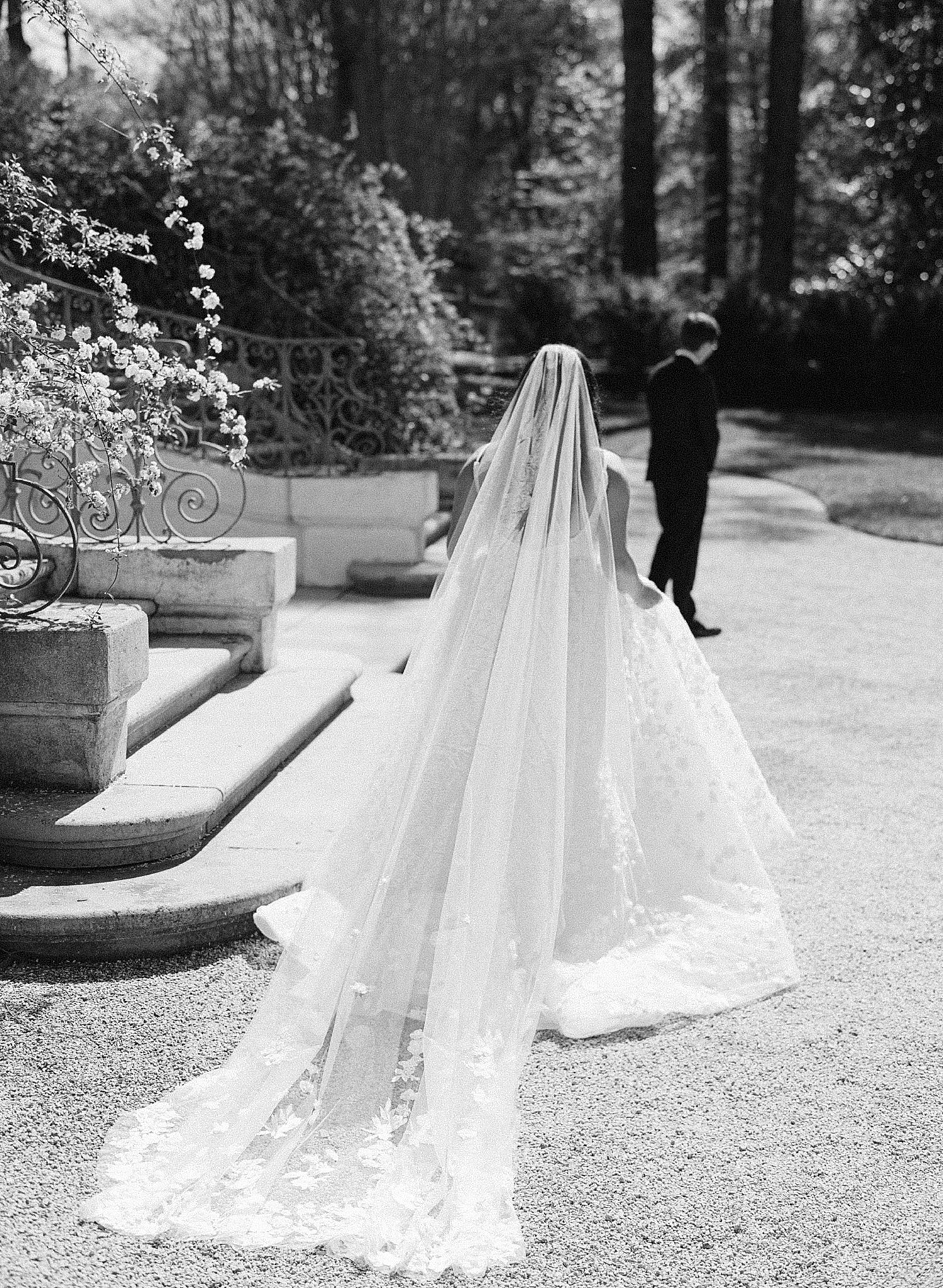 A black and white image of a bride walking towards her groom. Her veil trails on the ground behind her.