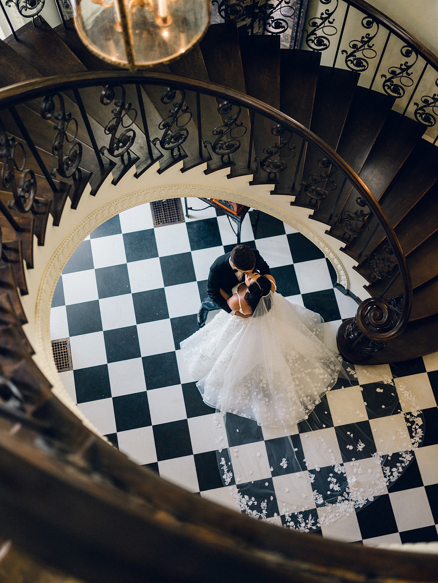 A top down view of a bride and groom dancing under a spiral staircase with checkered flooring underneath