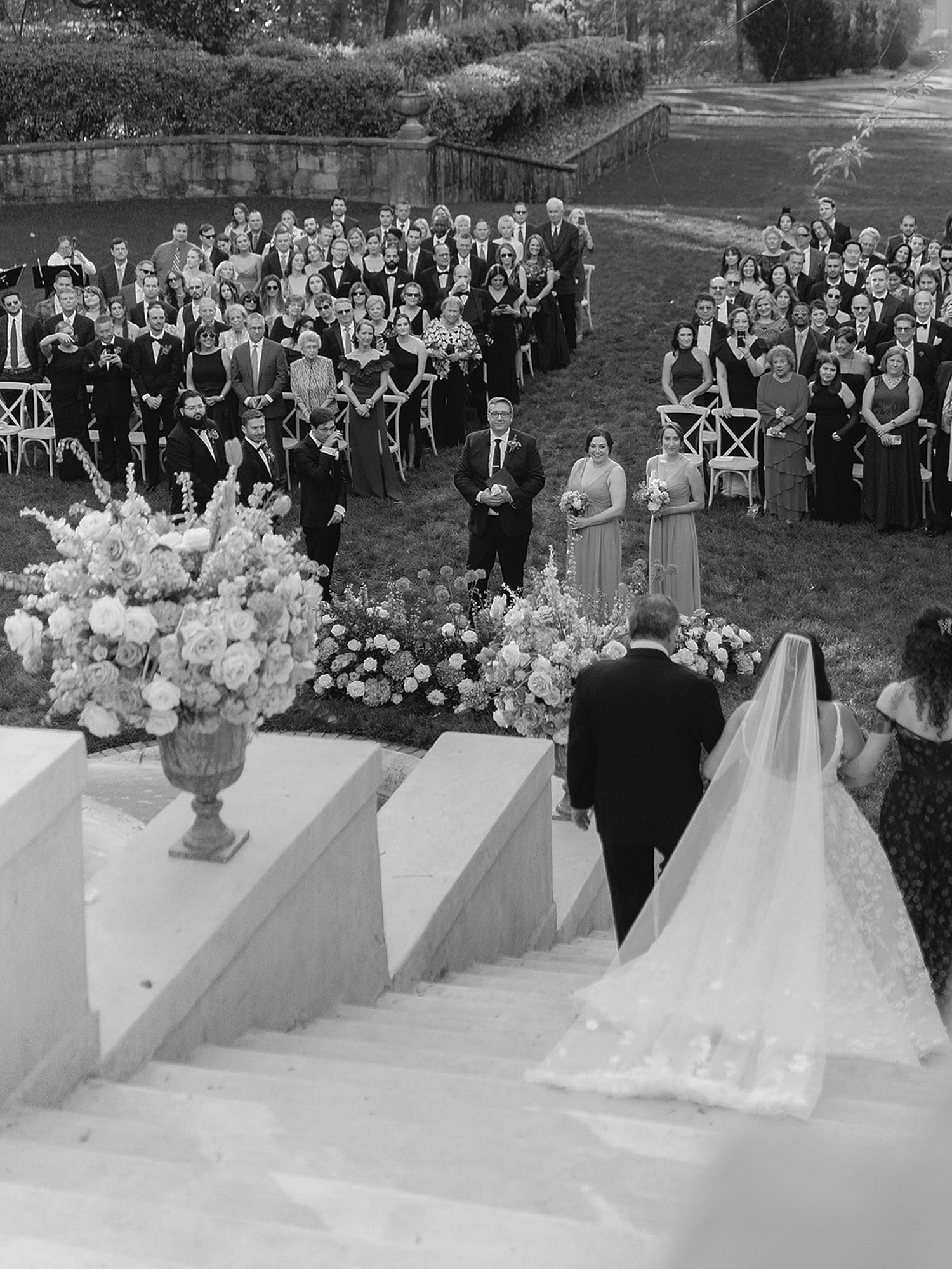 A black and white image of a bride walking down the Swan House stairs with her parents escorting her. The photo is taken from behind and you can see the groom's reaction in the background.