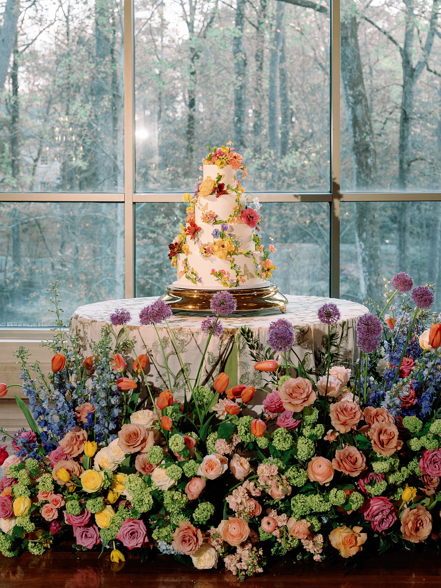 Wedding cake with four tiers and adorned in flowers and butterflies. Wild, colorful floral arrangements are on the ground in front of it, framing the cake from below.