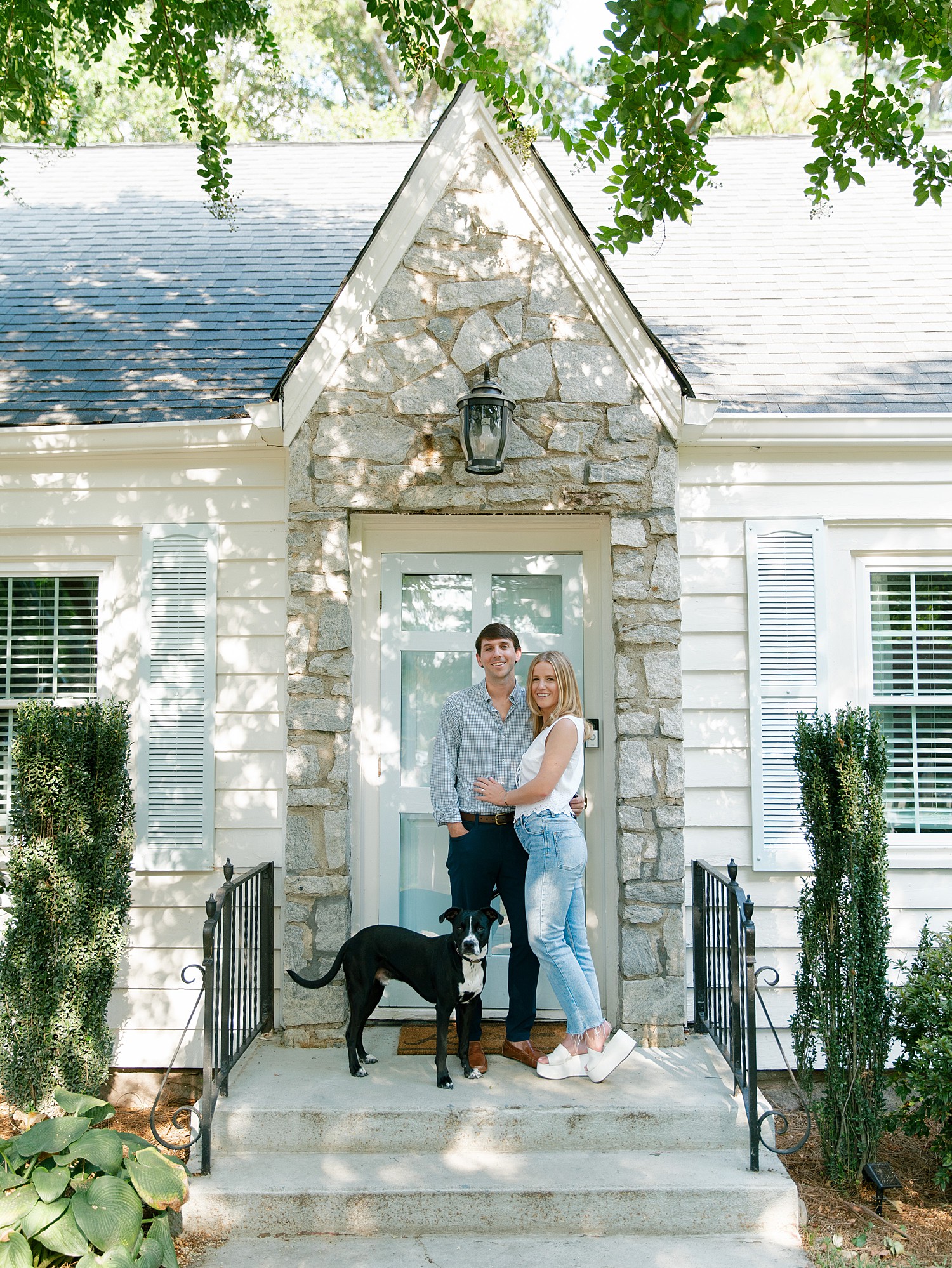 A couple smiles with their dog at the front door in Atlanta. Their door is light blue and their home has a white washed gray stone entrance with white siding