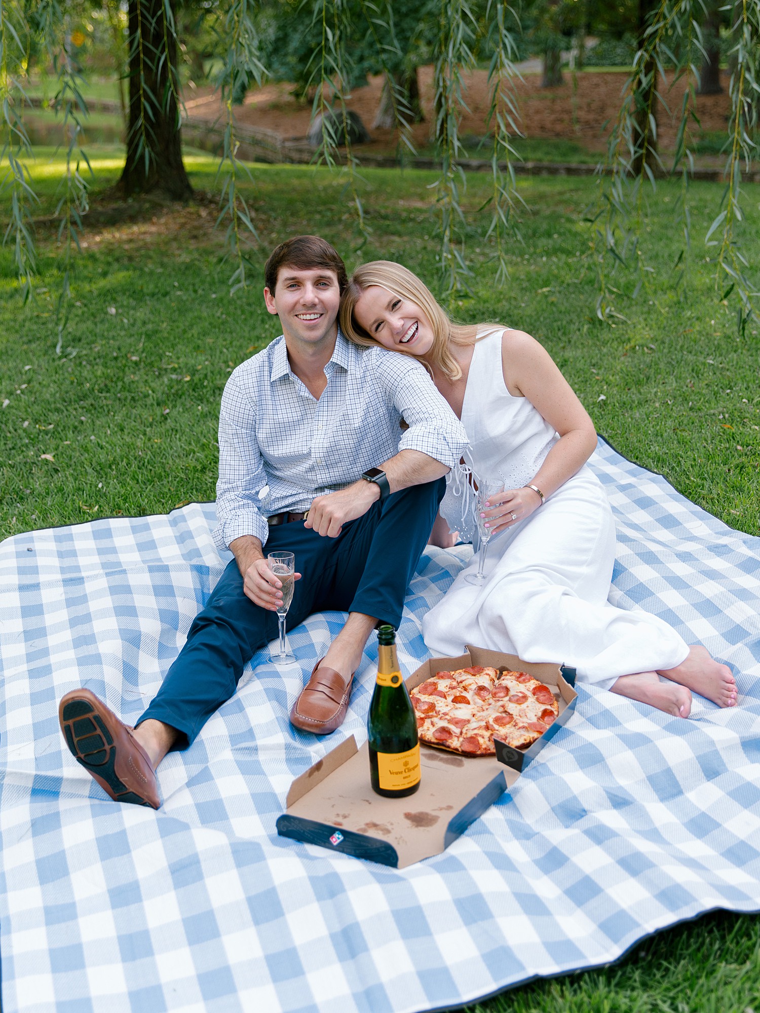 A couple smiles while sitting on a blue and white checkered picnic blanket with a box of Dominos pizza and a glass of Veuve Clicquot on the blanket next to them