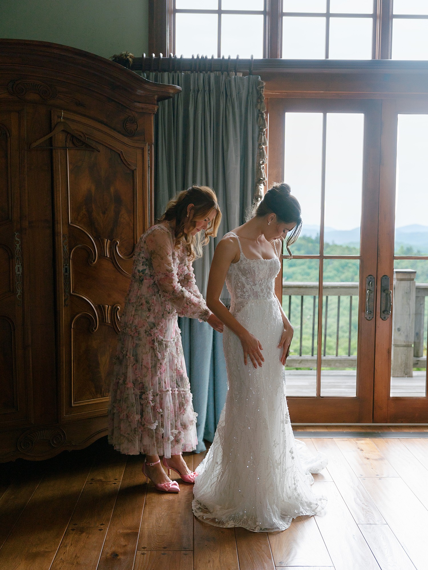 A mother of the bride buttons up the bride's dress in front of a wood armoir and a pair of glass doors that show the mountains in the background