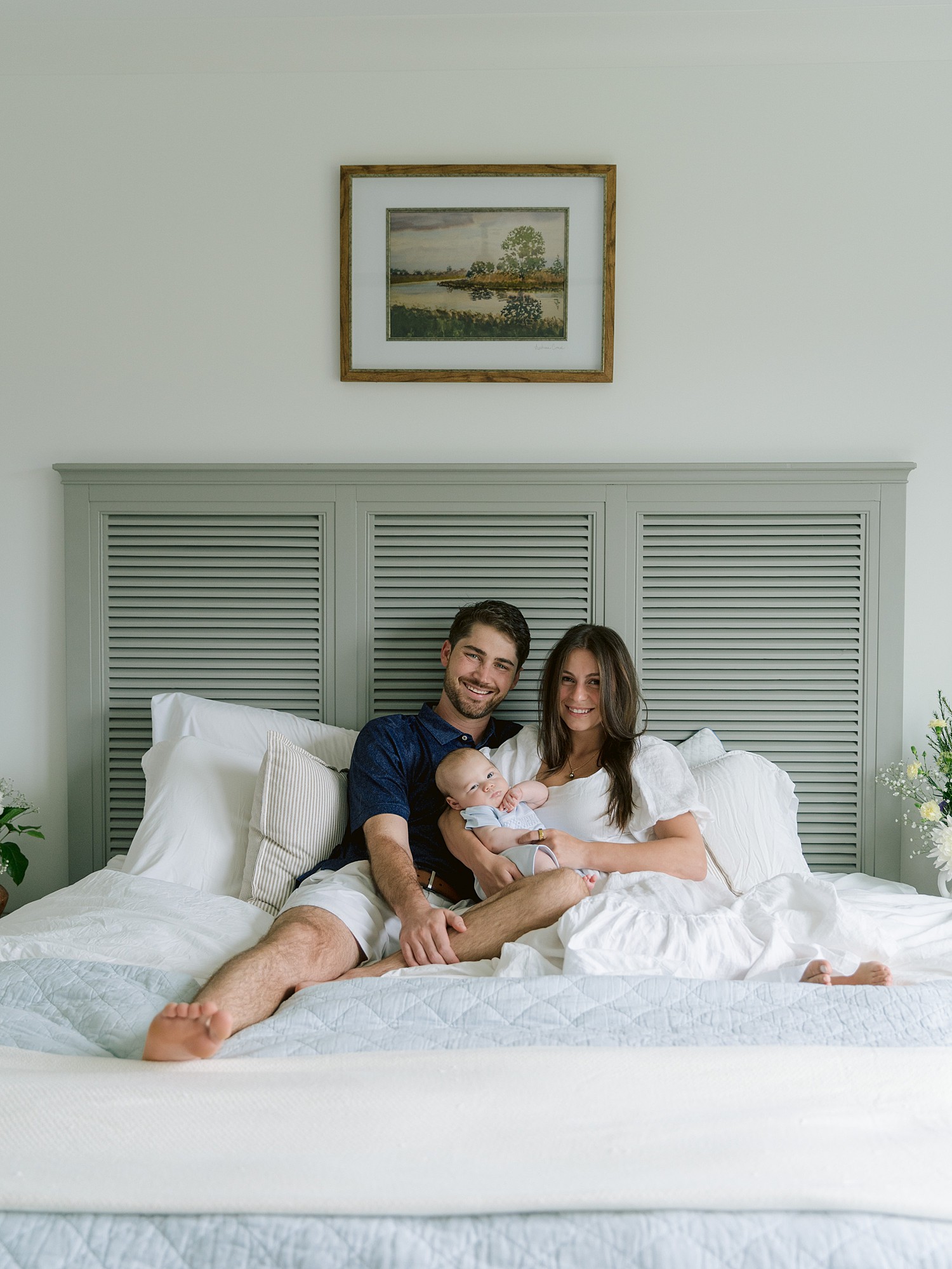 Family portrait of young parents with their new baby sitting on a bed with a landscape photo hanging above the bedframe