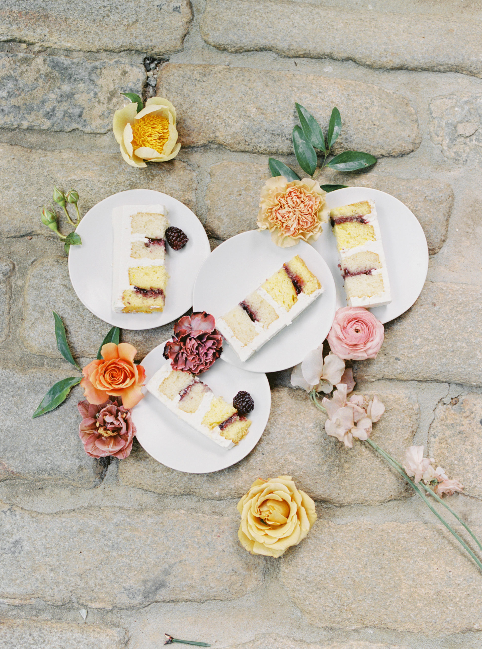 Slices of cake on white circular plates surrounded by flower stems on cobblestone