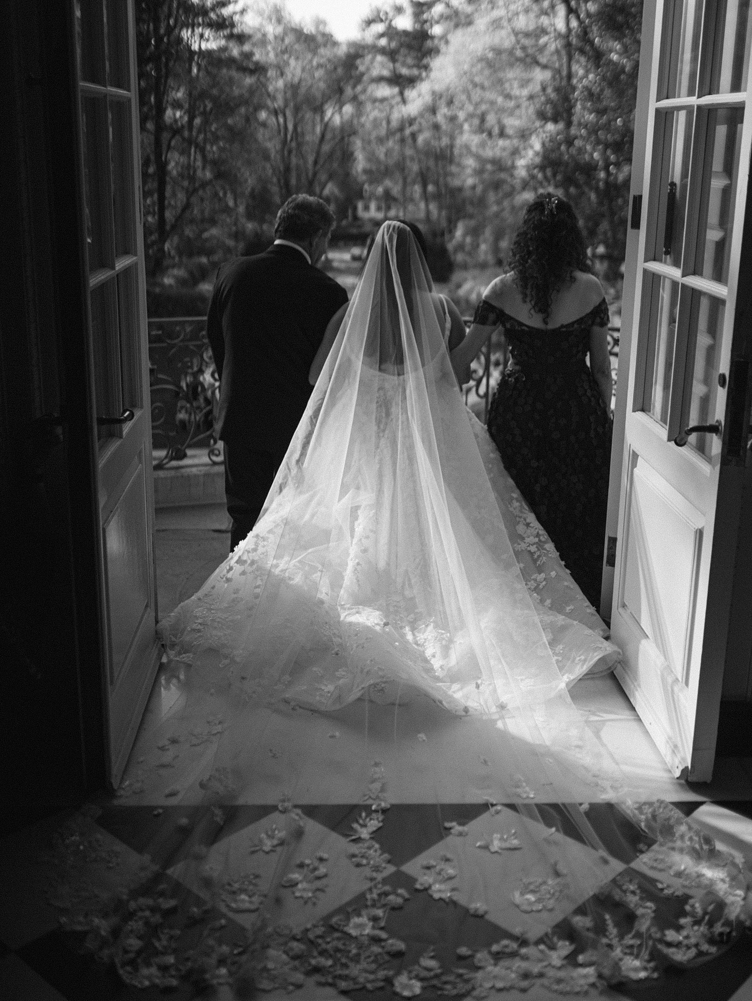 A black and white photo of the back of the bride's dress as she walks out the doors to her ceremony